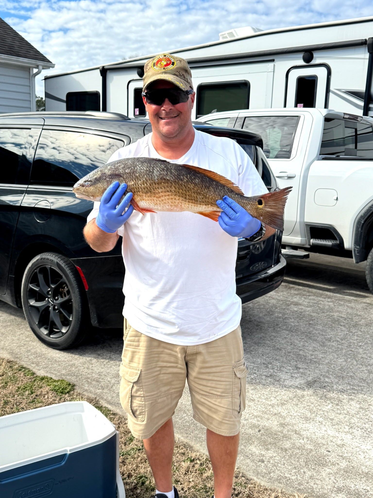 Man holding a large fish outdoors in a driveway, smiling in sunglasses and blue gloves.