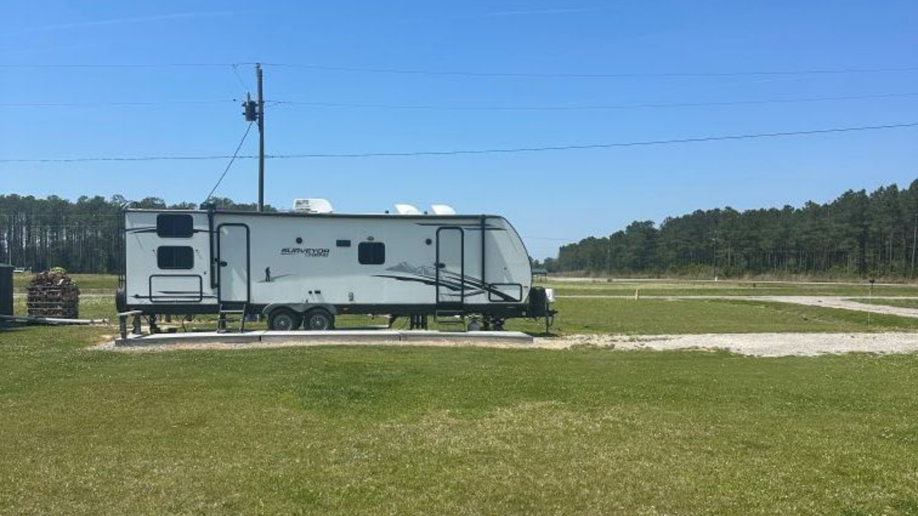 Aerial view of a lakeside campsite with RVs, cabins, and a dock beside grassy fields and forest.