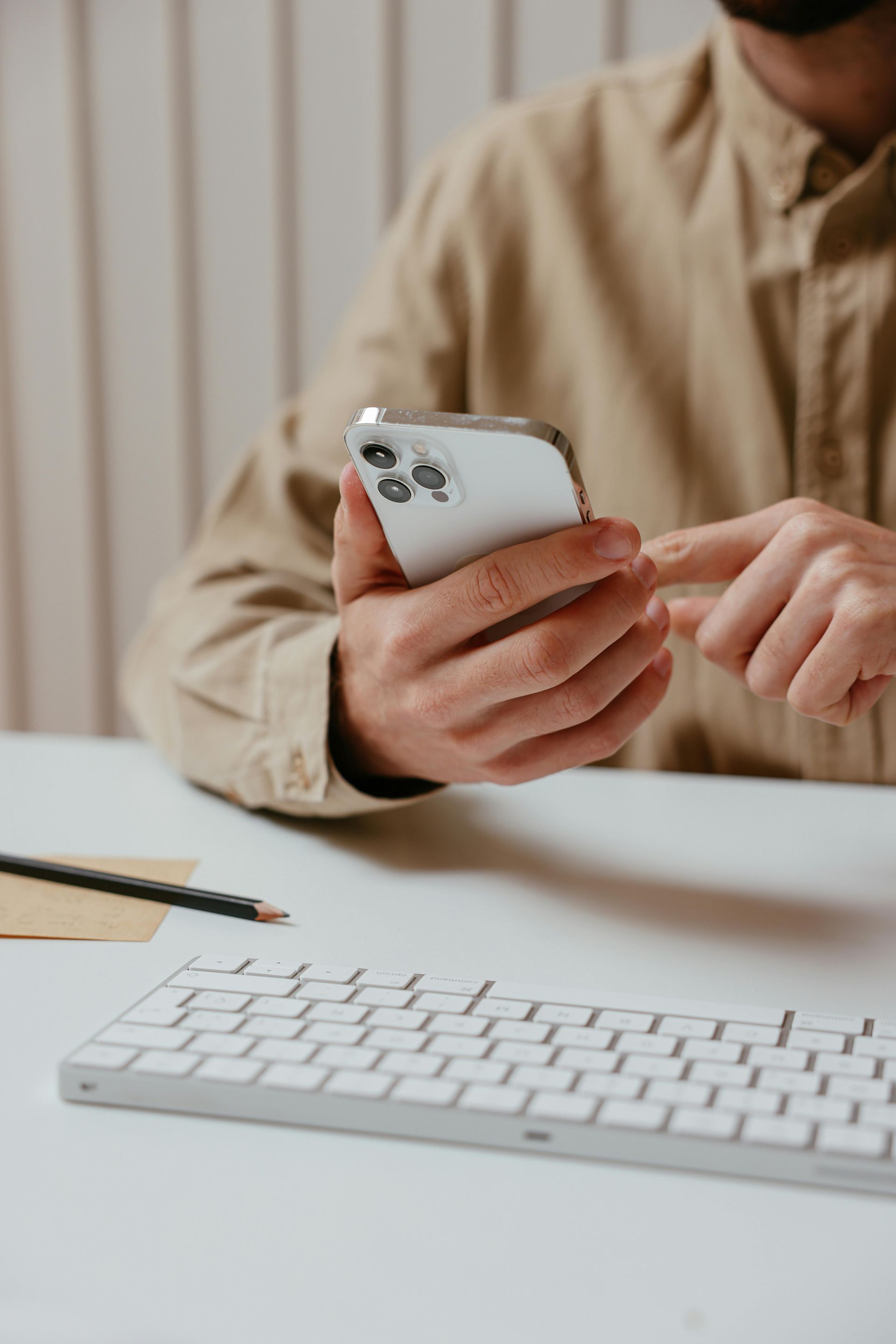 Person using a smartphone at a desk with a white keyboard and notebook.