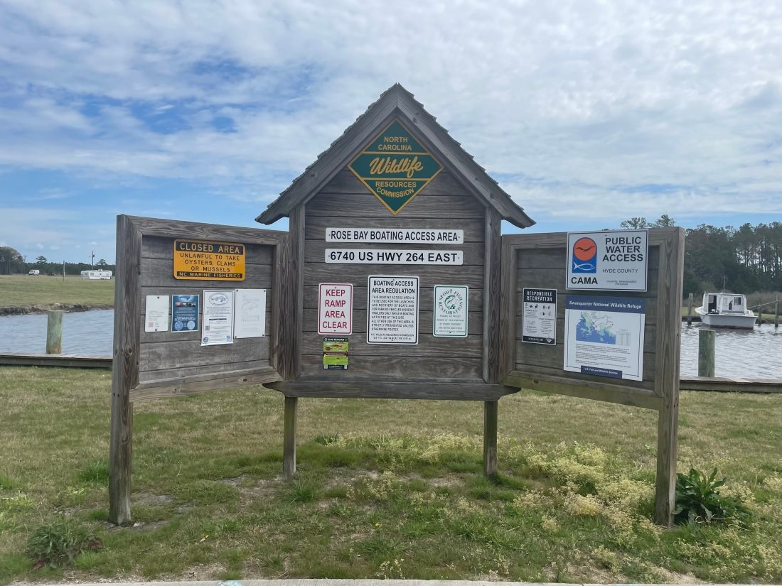 Community bulletin boards by a waterfront, with signs and notices on weathered wooden posts under a cloudy sky