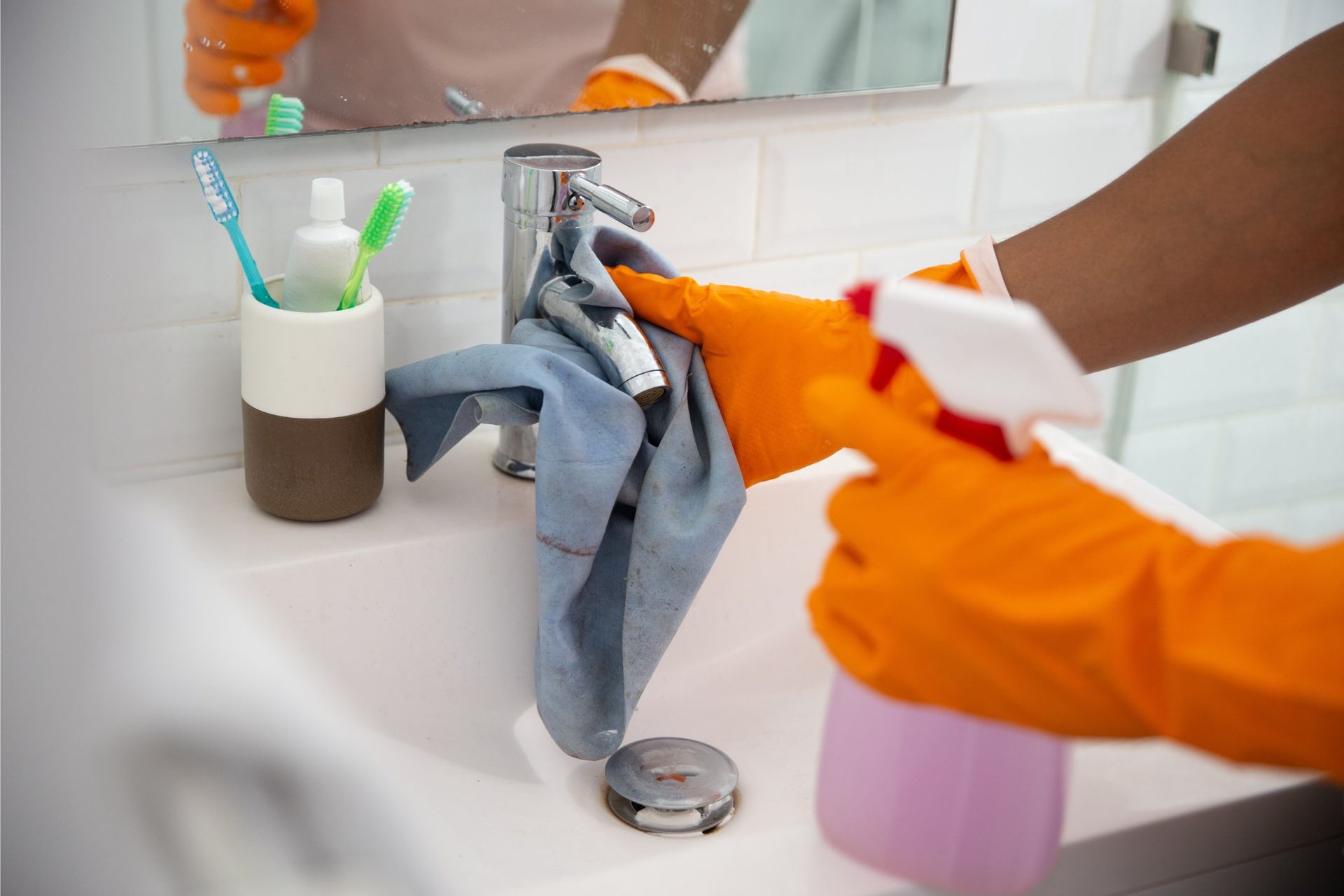 A person wearing orange rubber gloves wipes a bathroom faucet with a grey cloth while holding a spray bottle.