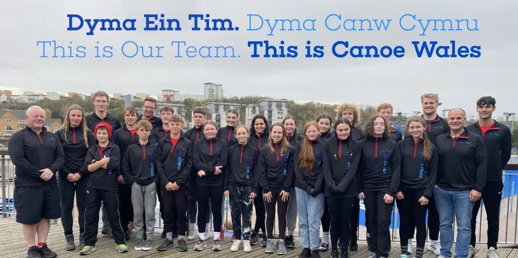 A group of people standing next to each other in front of a sign that says this is our team this is canoe wales.