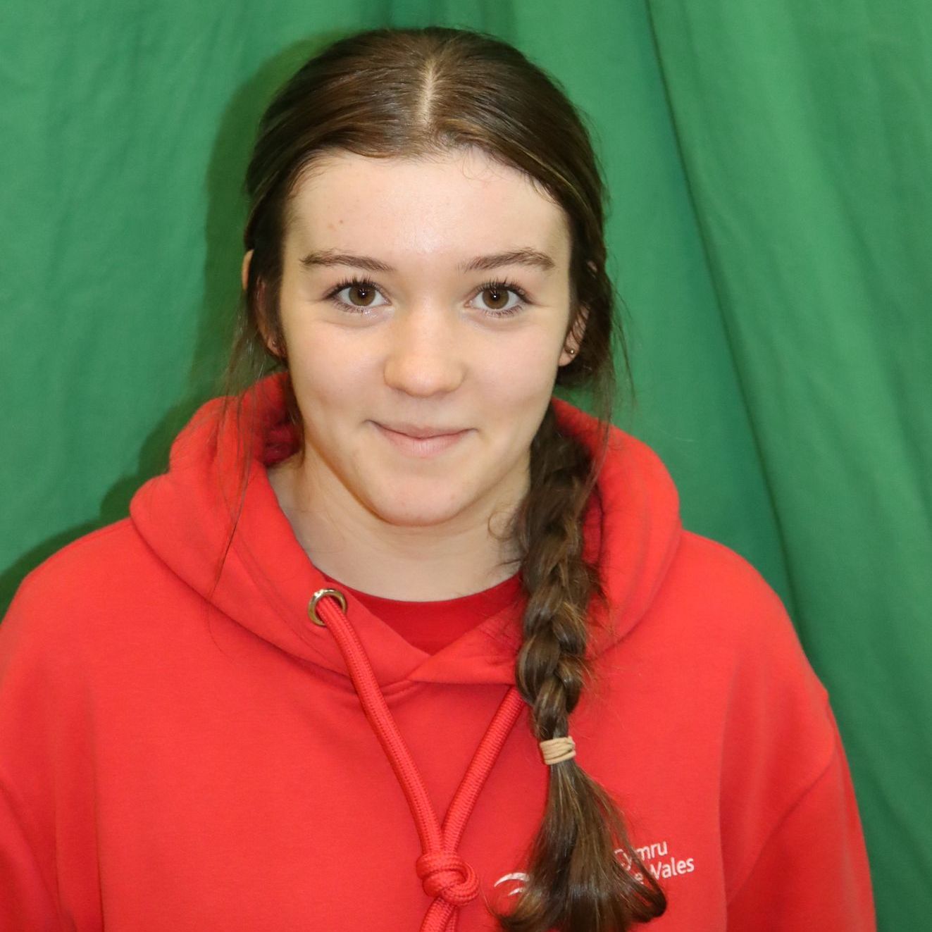 A young woman wearing a red hoodie with a braid is standing in front of a green curtain.