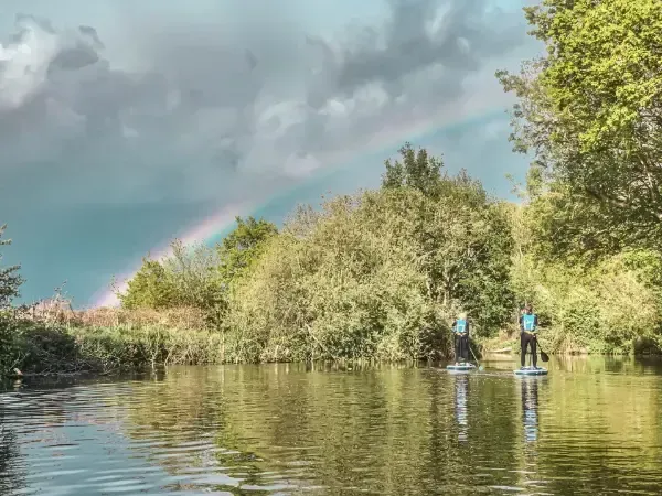 Two people are riding paddle boards on a river under a rainbow.