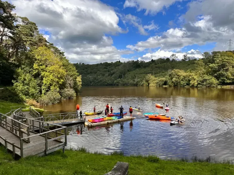 Mae grŵp o bobl yn sefyll ar doc mewn llyn.