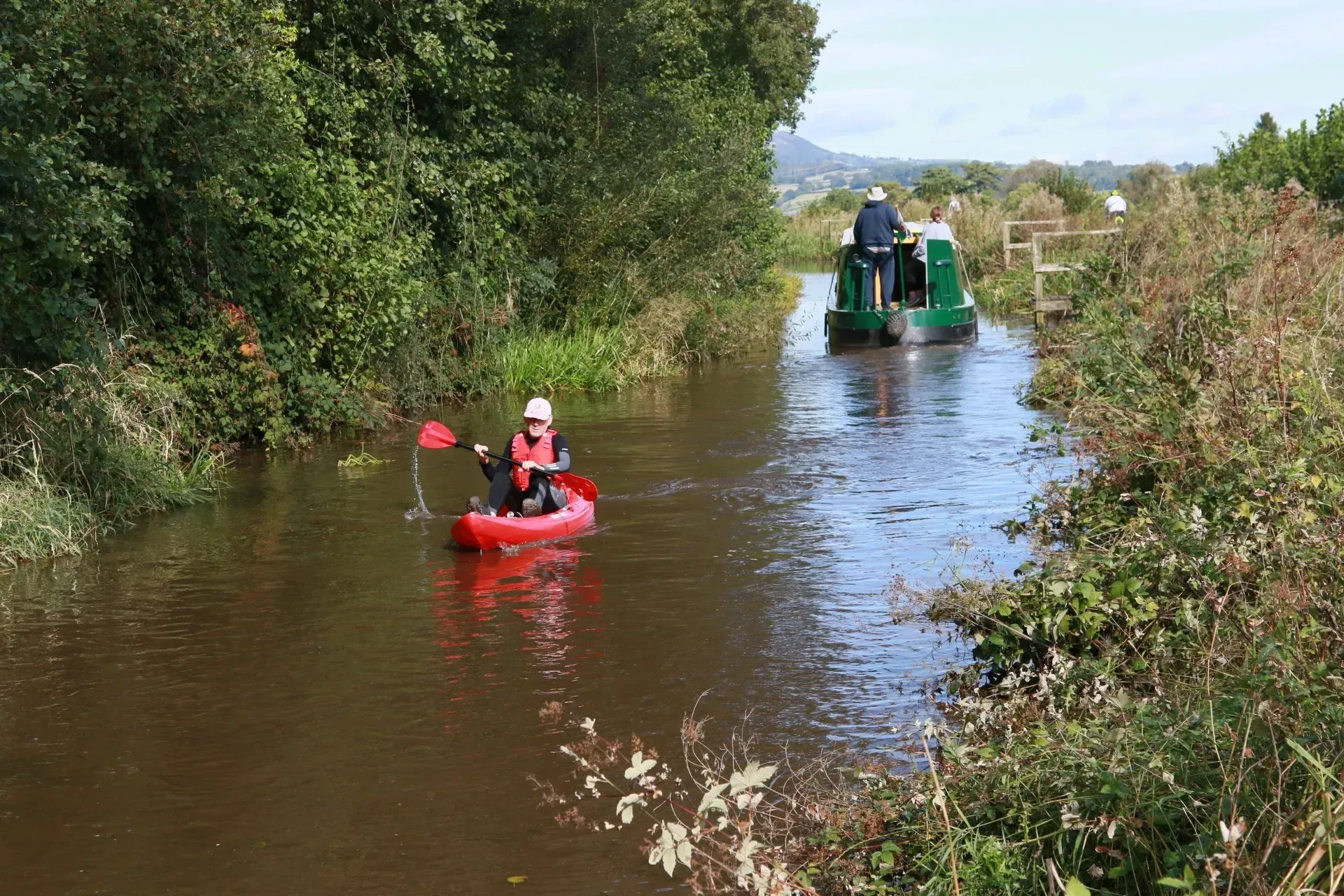 Mae dyn mewn caiac coch yn padlo i lawr afon wrth ymyl cwch gwyrdd.