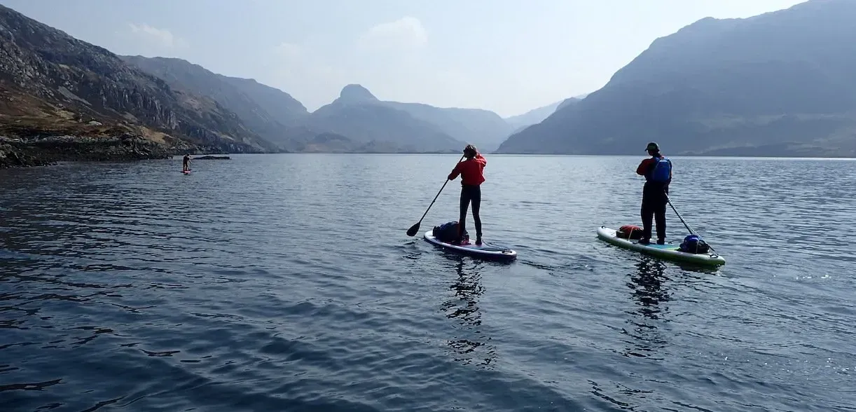 Two people are paddle boarding on a lake with mountains in the background.