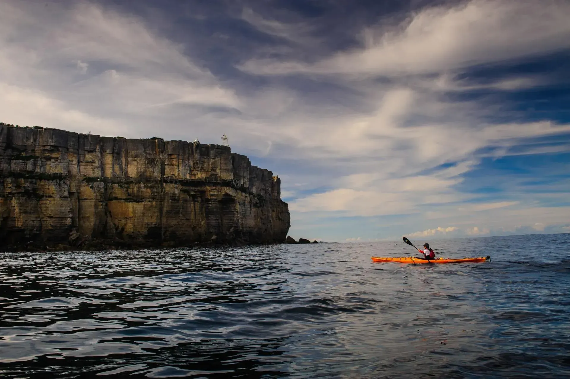 A person is paddling a kayak in the ocean near a cliff.