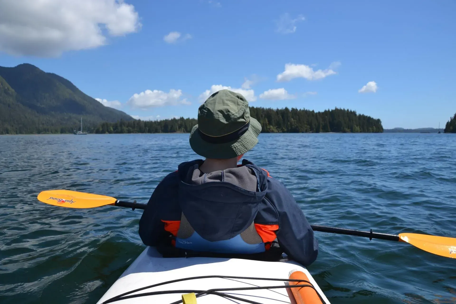 A person is paddling a kayak on a lake with mountains in the background.