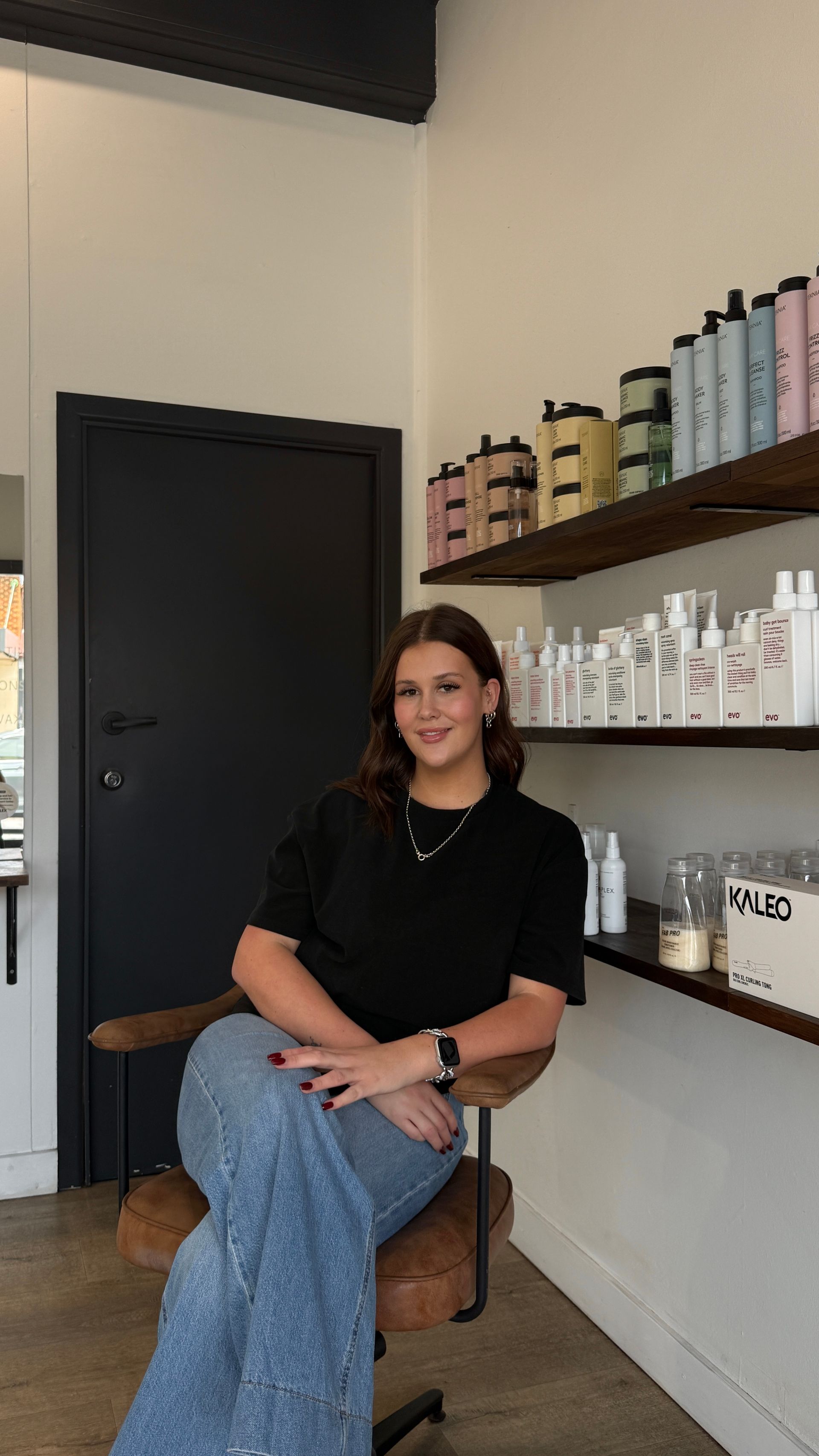 Woman seated in a salon chair, smiling. She's in a white top, shorts, and sandals. Sinks and products are behind her.