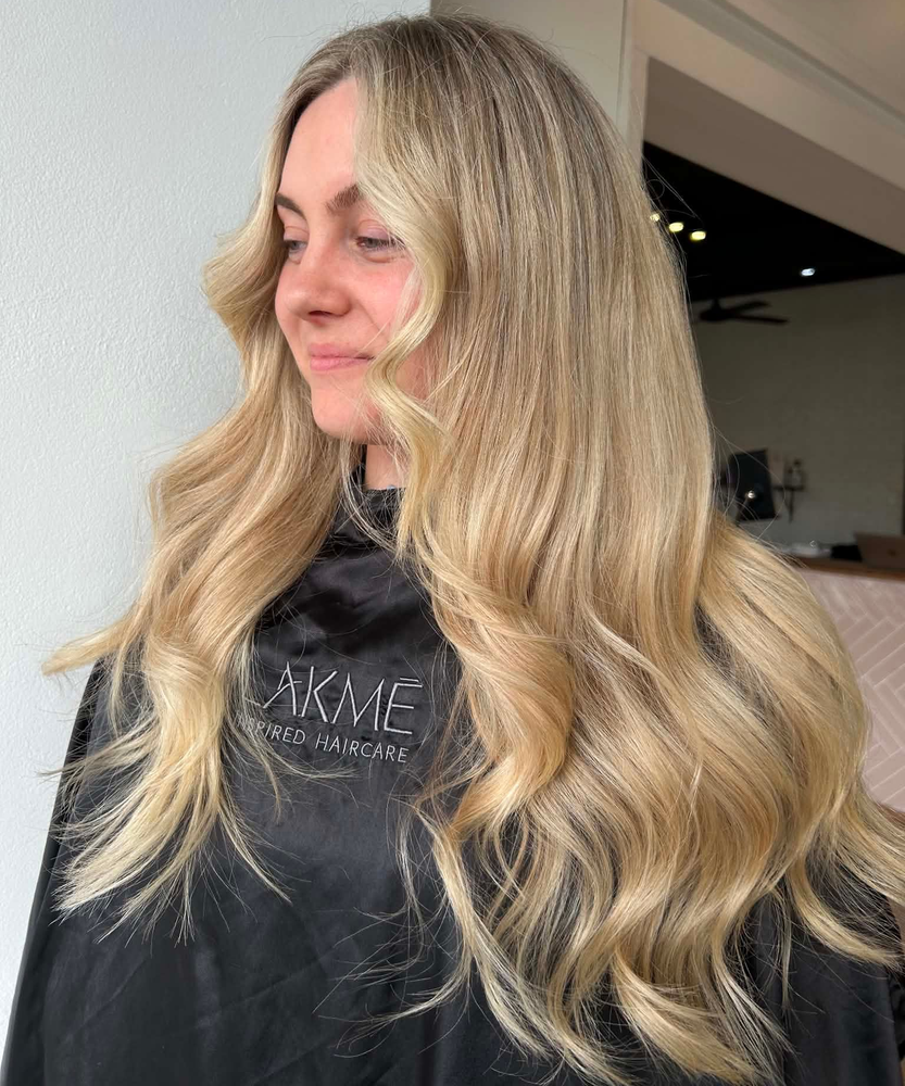 Woman with long, layered, light brown hair. She wears a gray sweater, in a salon setting.