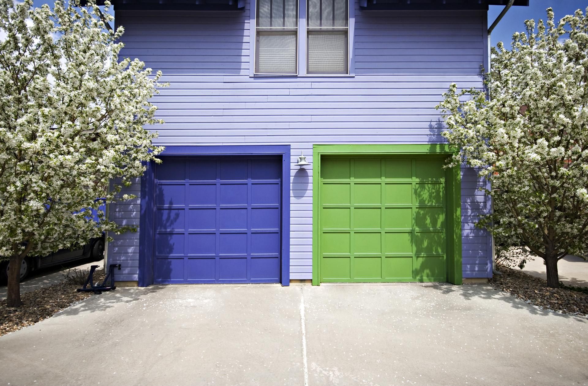 Two garage doors, blue and green, beneath a purple house, flanked by flowering trees.