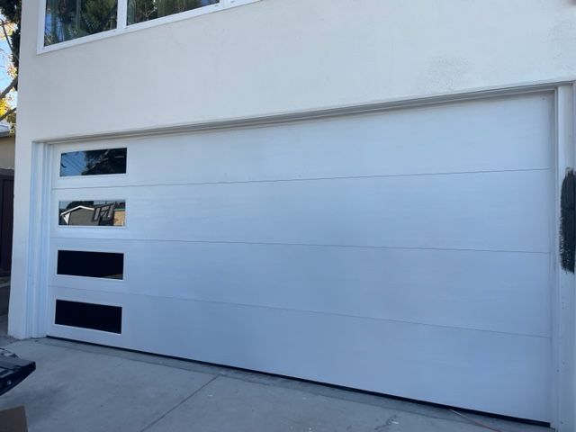 White garage door with three horizontal black windows and horizontal paneling; next to a white house.