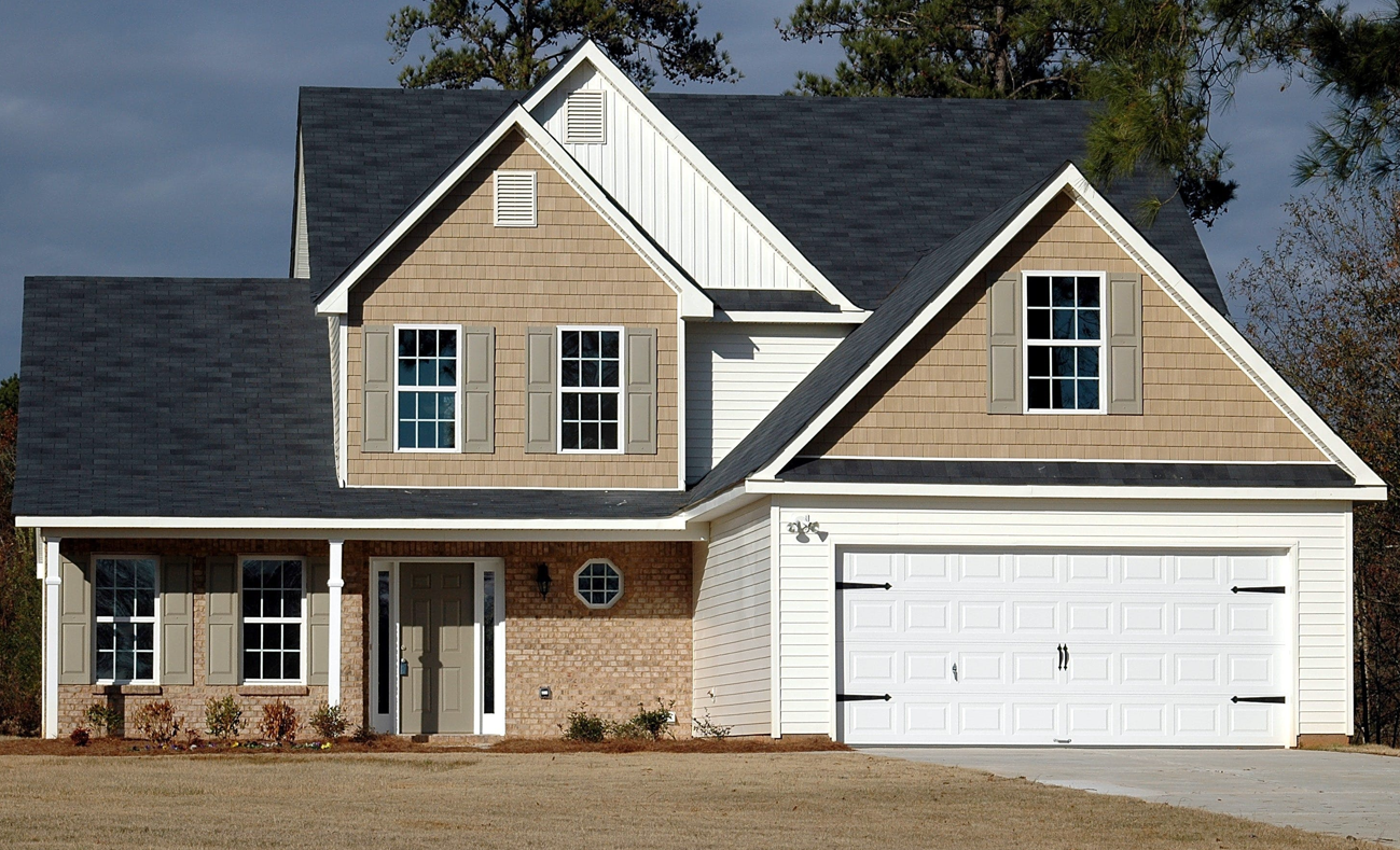Two-story house with a dark gray roof, tan siding, brick accents, and a white garage door.
