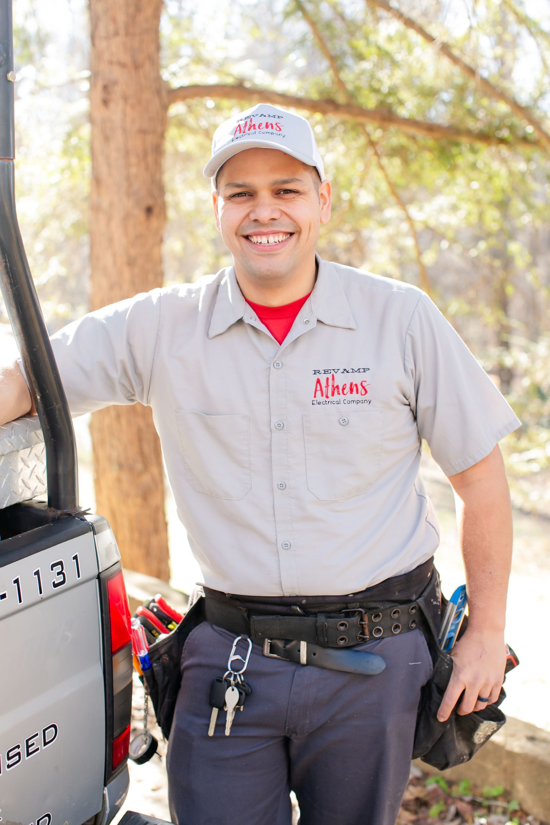 Smiling worker in a gray uniform and cap standing beside a vehicle in a wooded area.