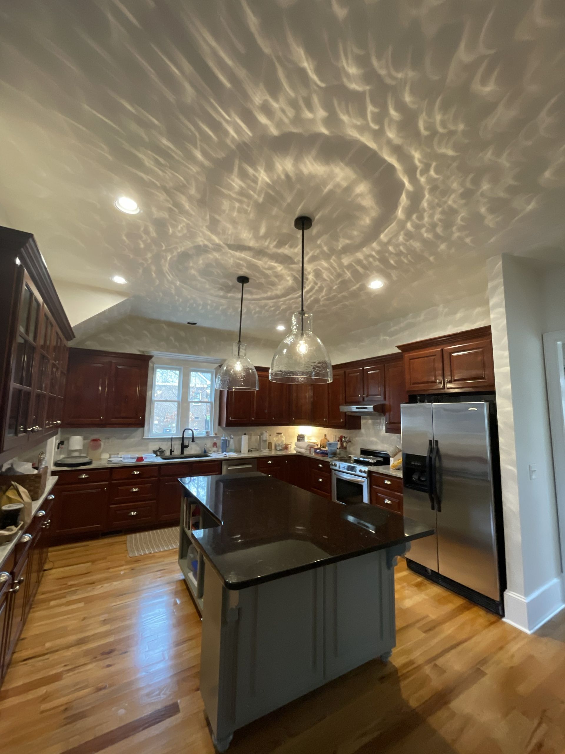Modern kitchen with dark wood cabinets, black island, stainless fridge, and circular light patterns on the ceiling