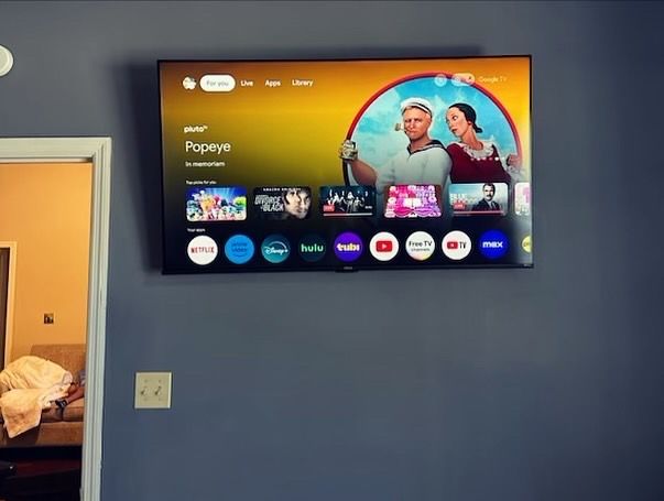 Wall-mounted TV on a blue wall showing a colorful streaming home screen with two people in a circular thumbnail.