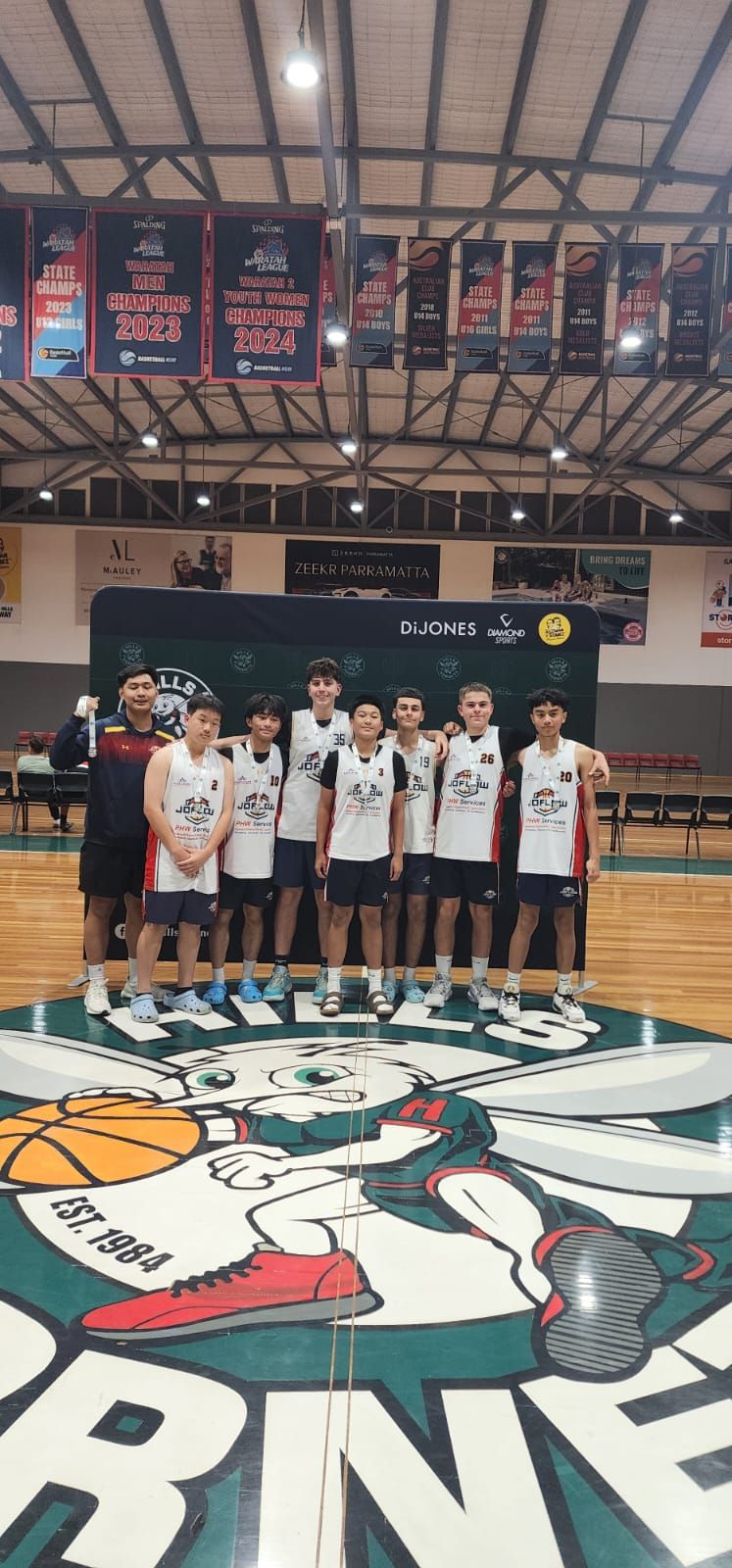 Basketball team poses on court with Hornets logo. Players in white jerseys. Indoor setting.