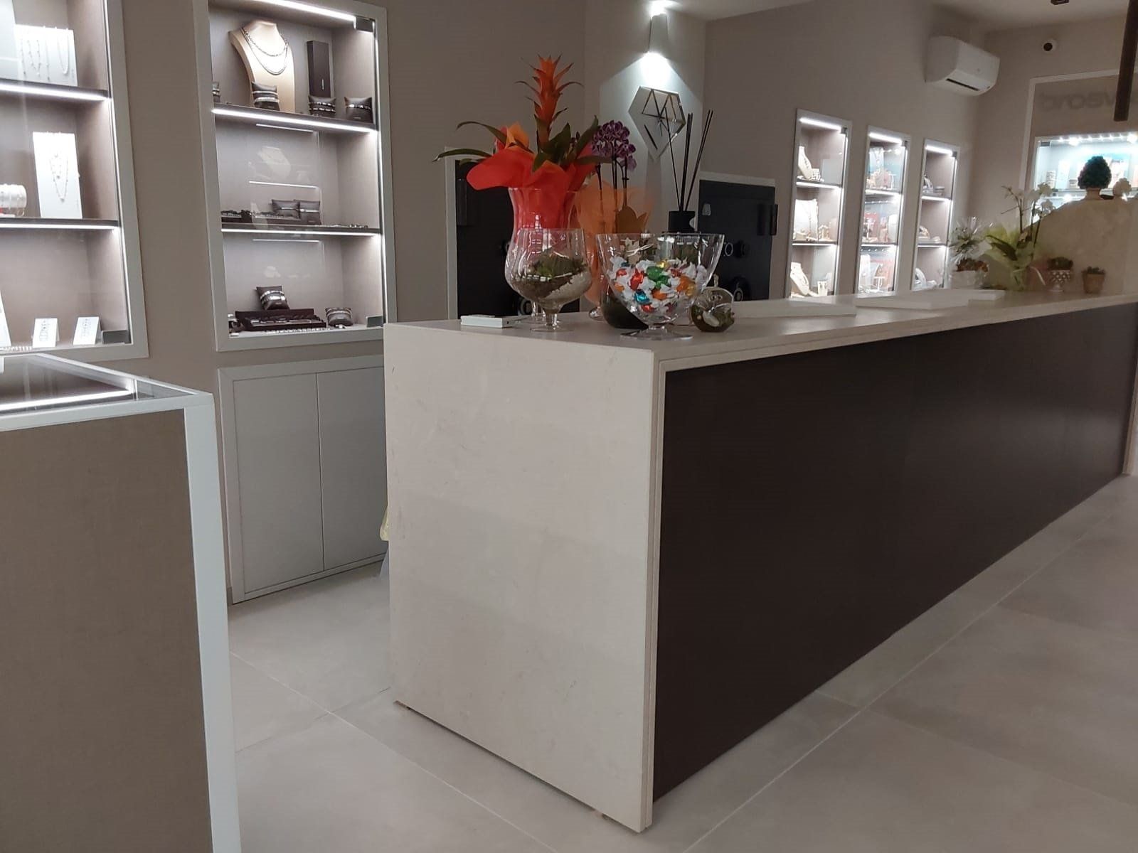Interno di una gioielleria con un lungo bancone bianco e marrone, vetrine e fiori.