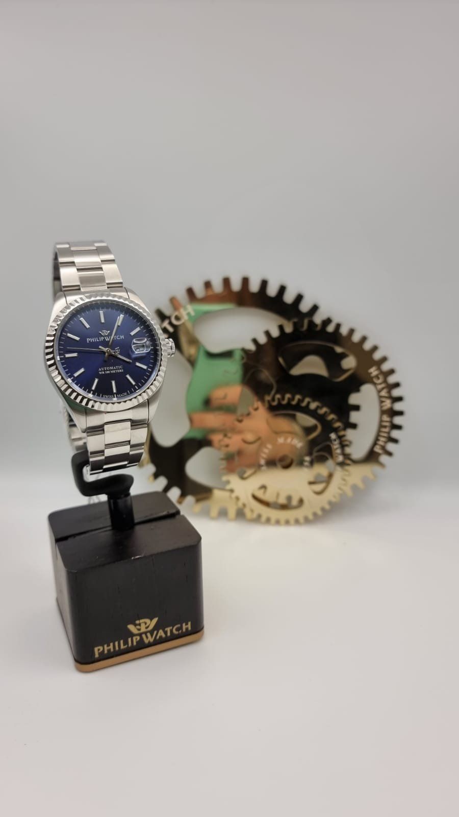 Orologio da polso in argento su supporto nero, accanto a ingranaggi decorativi su sfondo bianco.