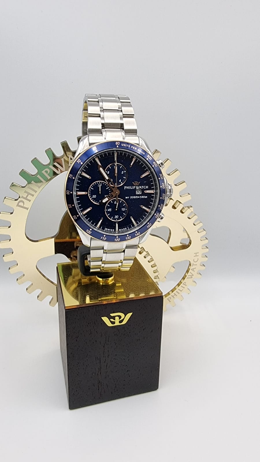 Orologio in argento con quadrante blu, su supporto nero e oro, con sfondo a ingranaggi.