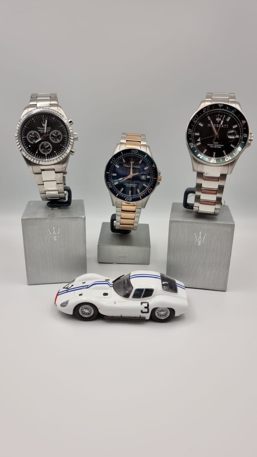 Orologi su supporti con un'auto da corsa giocattolo. Tre orologi, due argentati e uno dorato, con un'auto bianca e blu.