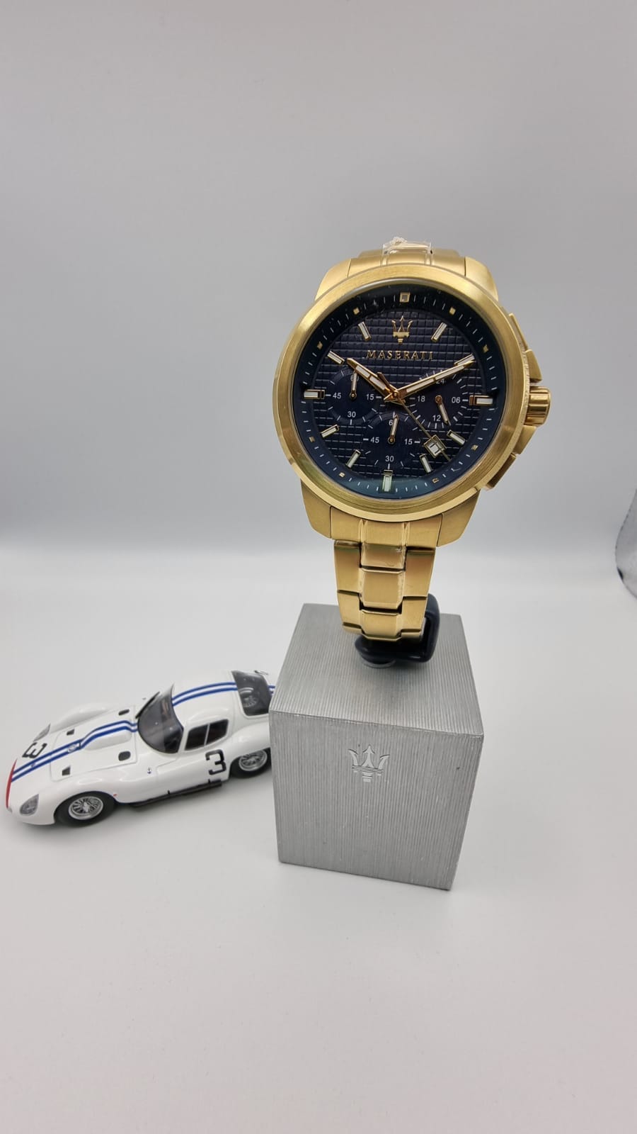 Orologio da polso in oro su un blocco grigio, accanto a un modello di auto da corsa bianca con strisce blu su sfondo bianco.