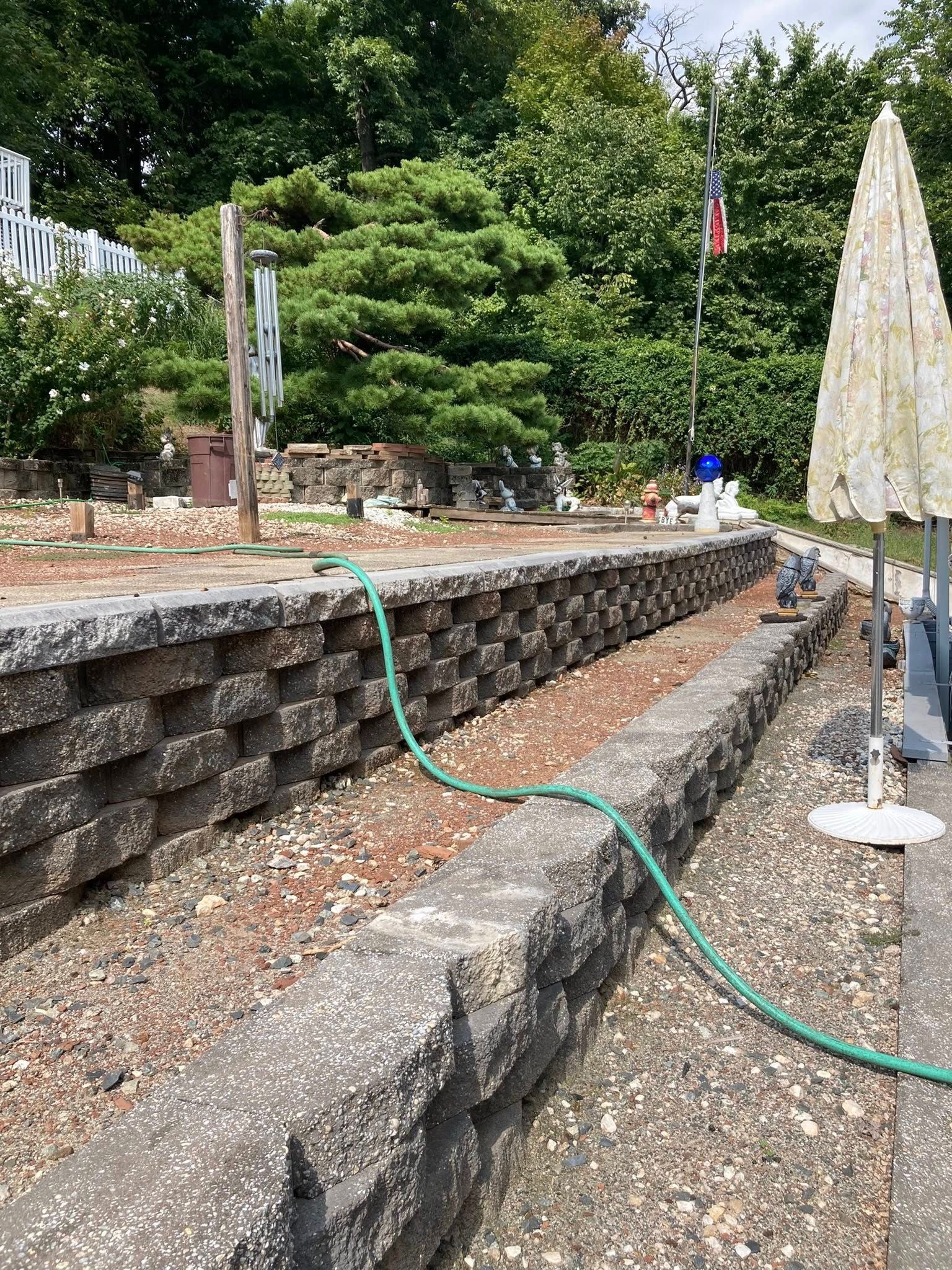 Stone retaining wall with a green hose, flag pole, and trees.