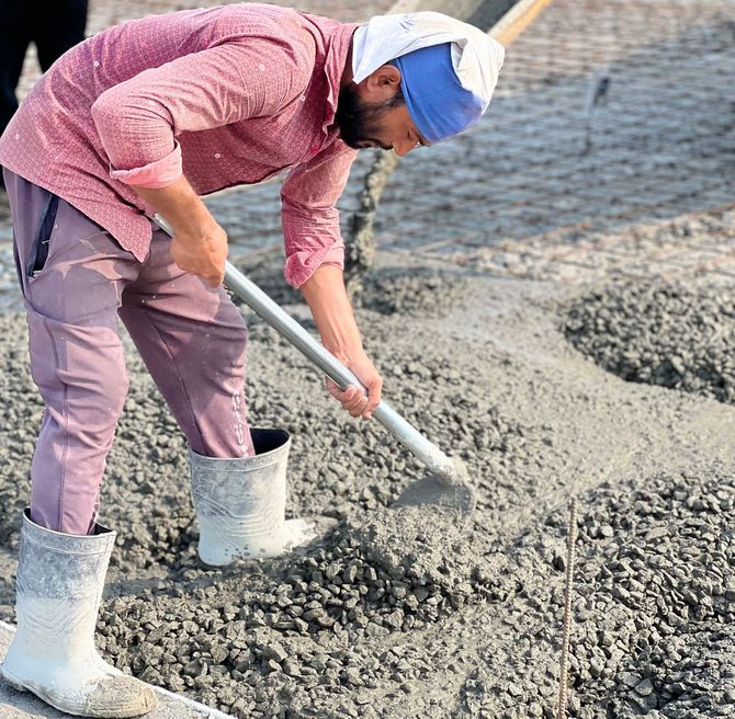 Worker in pink shirt and boots shoveling wet gravel at a construction site.