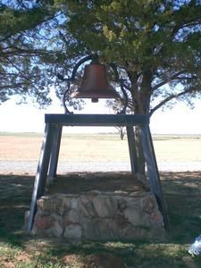 Bell hanging in a wooden frame over a stone base by a lakeside, with trees overhead