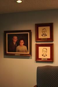Framed portraits on a beige wall in a dimly lit room, including one large photo and two smaller sketches.
