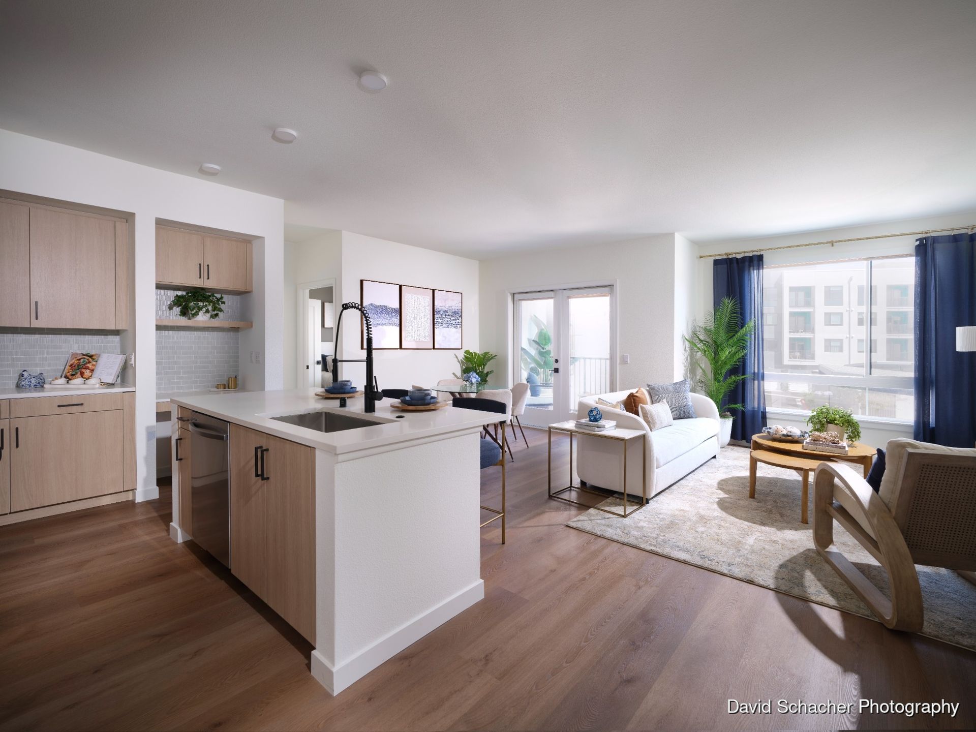 Modern living room with open kitchen, light wood floors, and neutral furnishings.