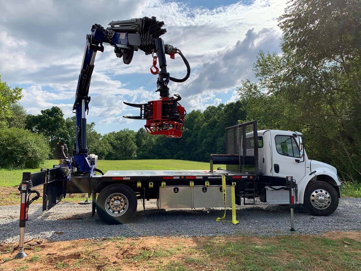 A white truck with a crane attachment, holding a red and black tree harvesting head, on a grassy field.