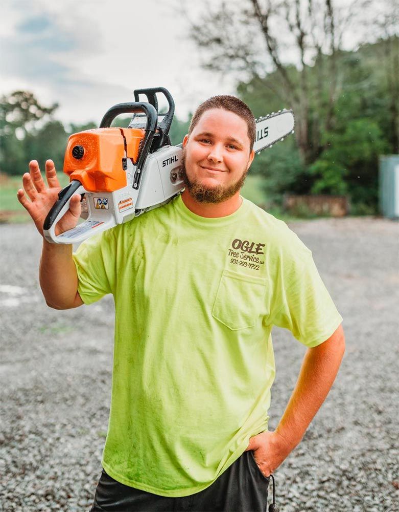 Man wearing a lime green shirt with a chainsaw on his shoulder, smiling. Outdoor setting.