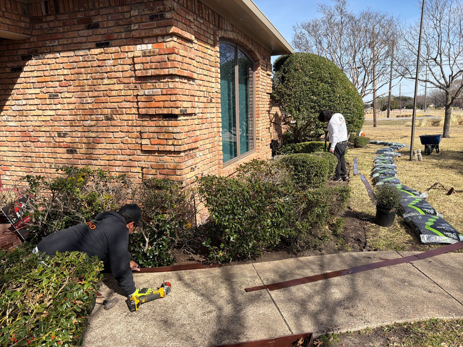 Two workers install edging along a residential garden bed next to a brick house.