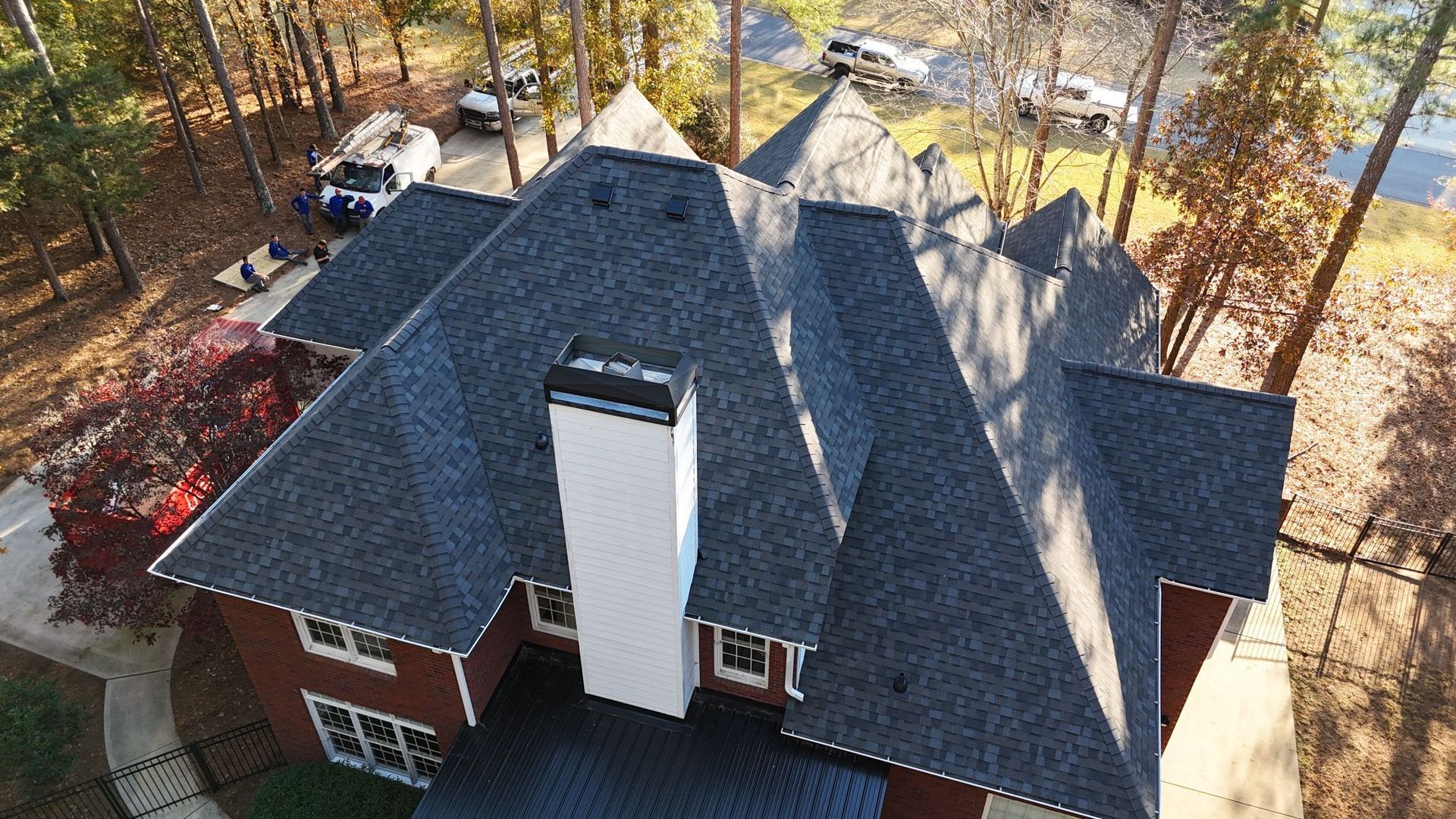 Aerial view of a dark shingled roof with a white chimney on a red brick house, with fall foliage surrounding.