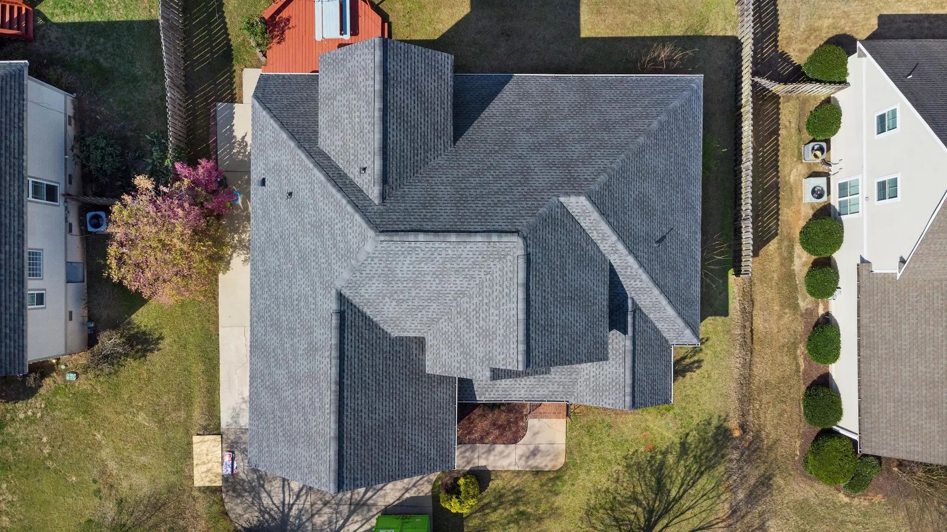 Overhead view of a gray roof on a house with a complex angular design surrounded by grass and neighboring buildings.
