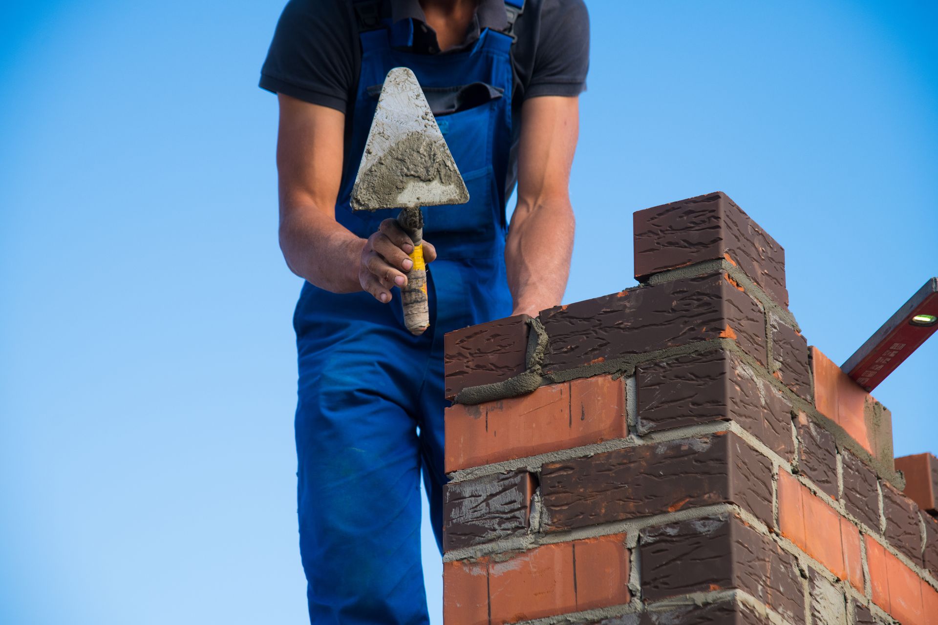 Bricklayer in blue overalls troweling mortar on a brick wall against a clear sky