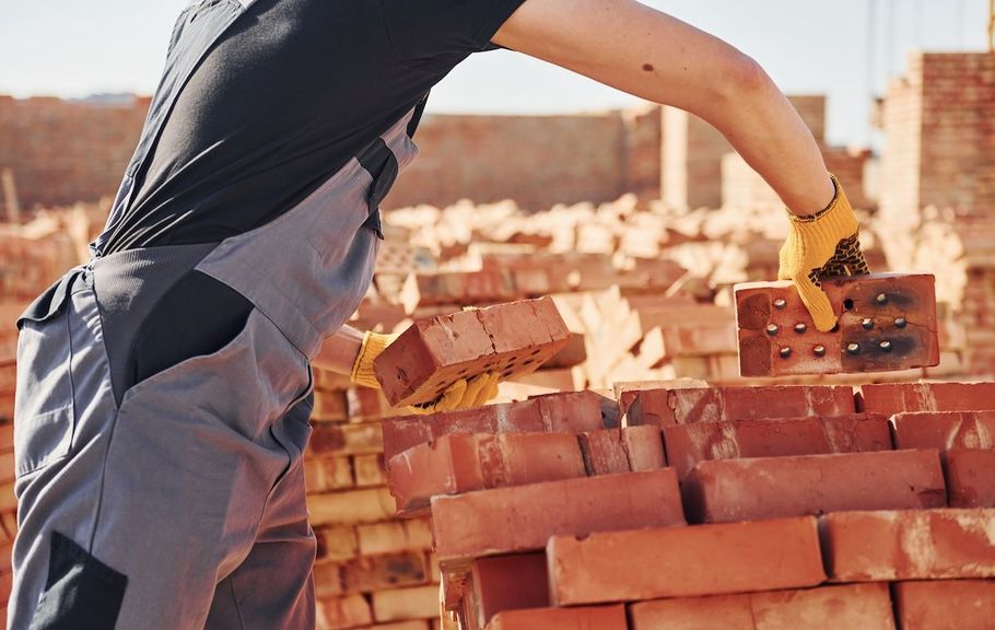 Worker stacking red bricks at a construction site, wearing gray overalls and yellow gloves.