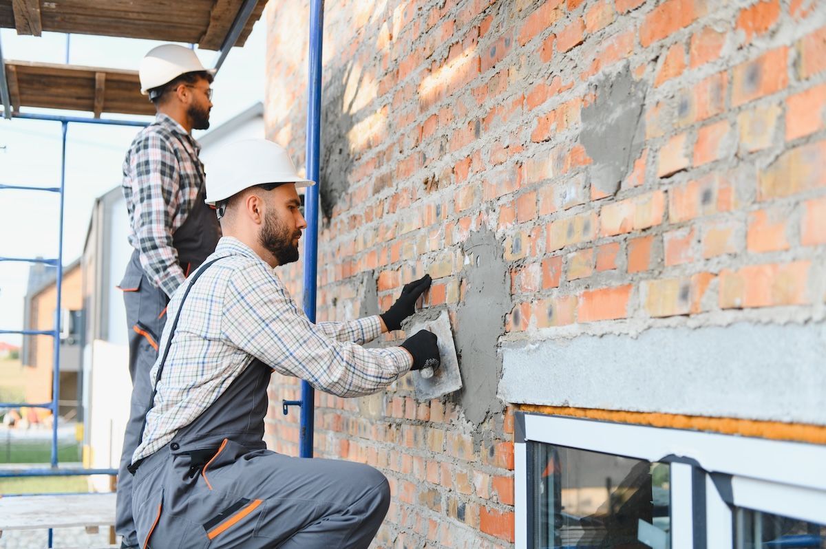 Two workers plastering an exterior brick wall on scaffolding