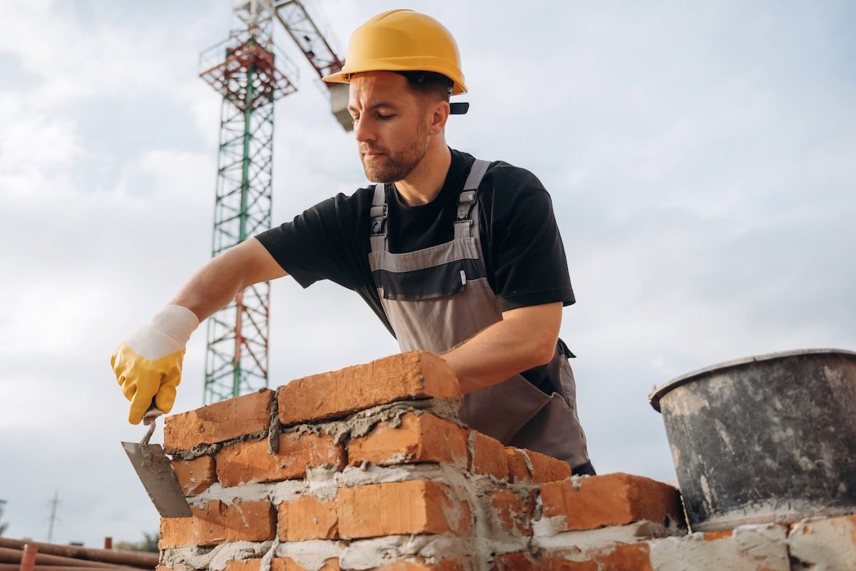 Construction worker laying bricks at a brick wall, wearing a yellow hard hat and gloves.