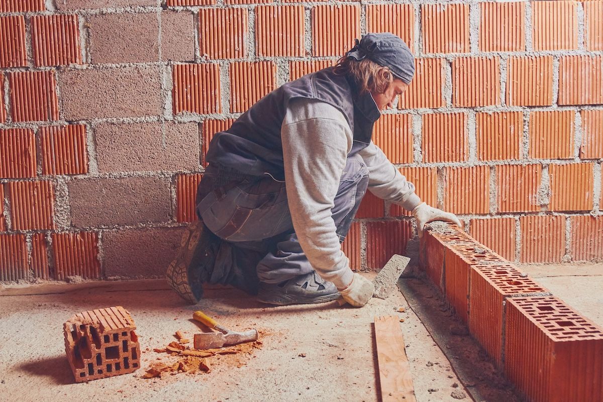 Mason kneeling on a construction site, laying bricks against a partially built wall.