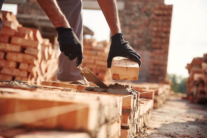 Gloved hands laying bricks with mortar at a construction site, with stacked bricks in the background