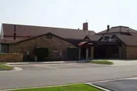 Single-story brick building with a sloped roof and covered entrance, viewed from across a parking lot.