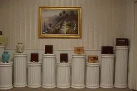 White pedestal display with assorted small objects beneath a framed painting in a beige room