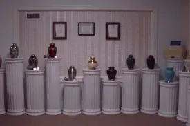 Row of urns on white pedestals in a room, with three framed pictures above them.