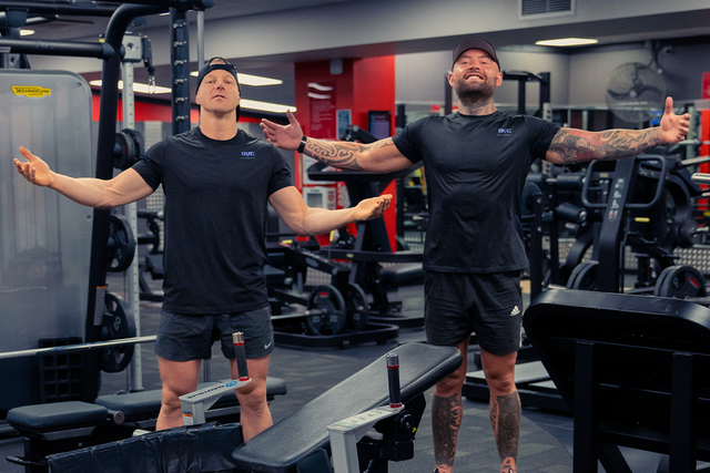 Two men are standing in a gym with their arms outstretched.
