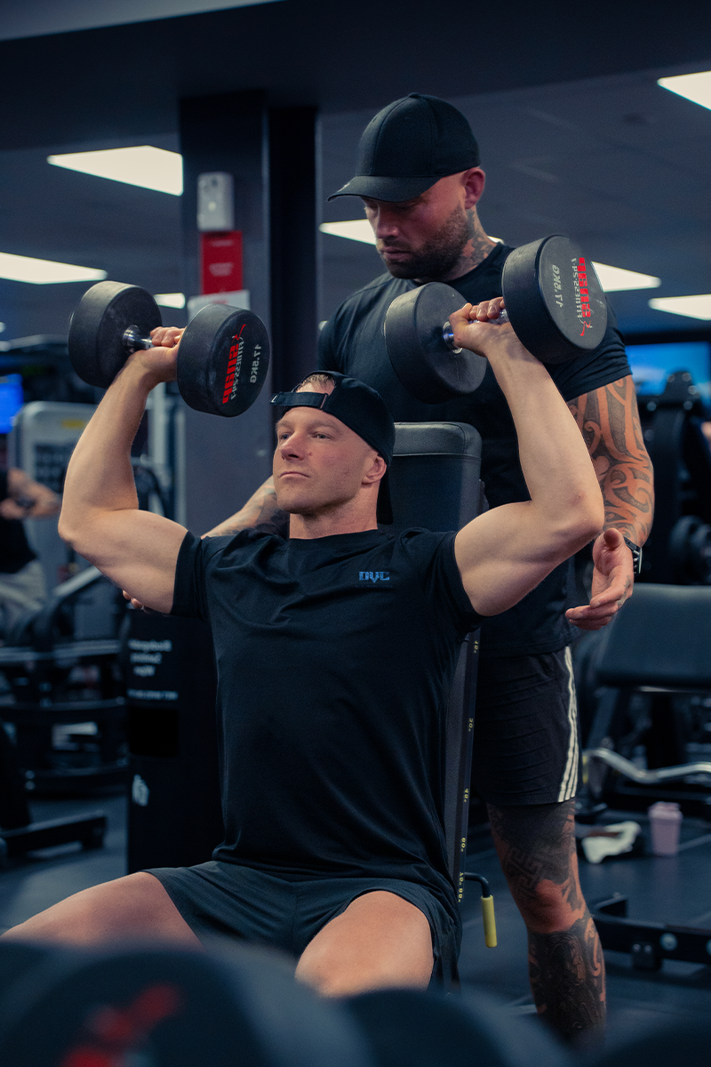 A man is lifting dumbbells in a gym while another man watches.