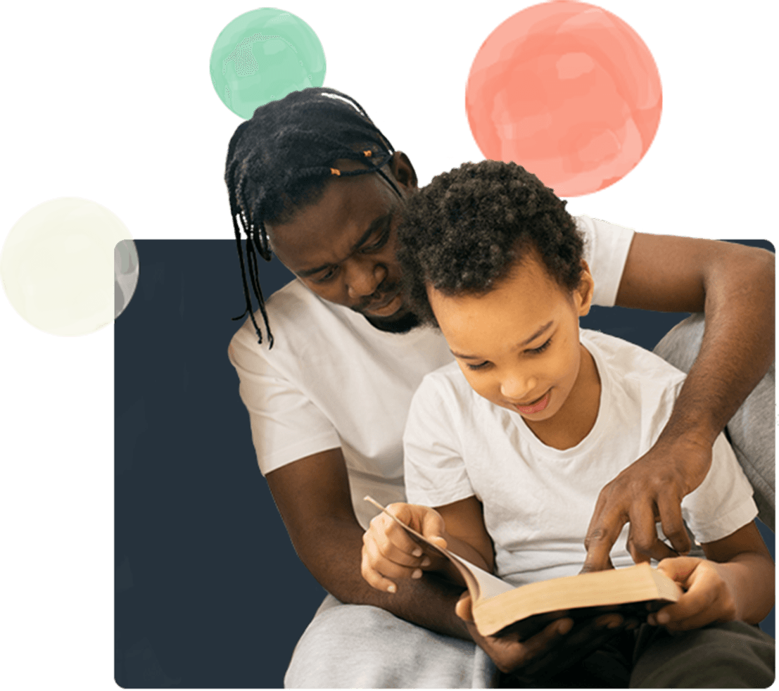 A father sits with his young son and helps him to read a book.