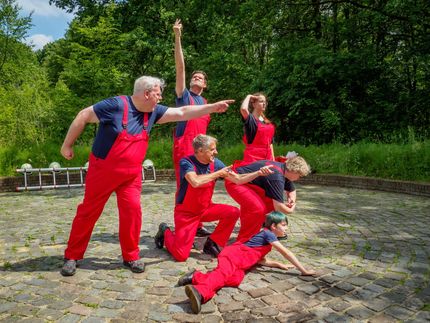 Five people in red overalls posing energetically outdoors on a stone patio, trees in the background
